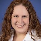 Headshot portrait of smiling woman with curly brown hair in white coat, blue background