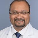 Studio headshot of smiling man in white medical coat and tie, light blue background.